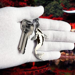 Può includere: Un portachiavi color argento con un ciondolo a forma di pesce koi. Il pesce ha segni bianchi e neri ed è attaccato a un portachiavi con due chiavi. Lo sfondo è sfocato, con toni rossi e verdi.