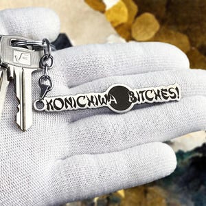 May include: Silver keychain with the text "KONICHIWA BITCHES!" in black lettering. The keychain is attached to a set of keys. The background is blurred.