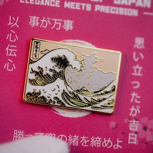 Op de afbeelding: Rechthoekige goudkleurige pin met een gestileerd golfontwerp in wit, zwart en beige, geïnspireerd op Japanse kunst. De pin heeft een gouden rand en staat op een roze achtergrond met Japanse tekst.