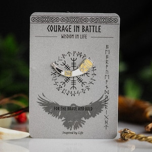 Op de afbeelding: Een Viking hoornspeld met zilveren en gouden details, gepresenteerd op een grijze kaart. De kaart is bedrukt met de woorden "COURAGE IN BATTLE, WISDOM IN LIFE" en "FOR THE BRAVE AND BOLD". Een ravenafbeelding staat onderaan.