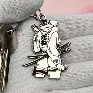 May include: Silver-colored keychain featuring a stylized anime character with two swords. The character is wearing a hoodie with Japanese text on the back. The keychain is attached to a key ring and a key.