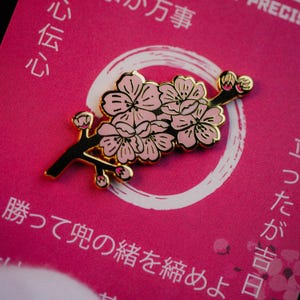 Könnte beinhalten: Ein rosa-goldener Emaille-Pin mit einem Kirschblütenzweig. Der Pin hat einen schwarzen Stiel und goldene Konturen, mit rosa Blüten und Knospen. Auf rosa Hintergrund mit japanischem Text.