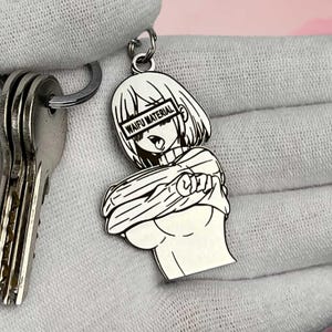 May include: Silver-colored anime-style keychain featuring a character with the text "WAIFU MATERIAL". The keychain is attached to a keyring with keys. The character is depicted with short hair and a sweater.