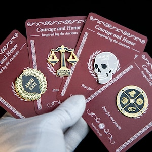Pode incluir: Uma coleção de pins em cartões bordô. Os pins apresentam designs como uma balança, uma caveira e um emblema circular com símbolos. Os cartões têm o texto "Courage and Honor" e "Inspired by the Ancients".