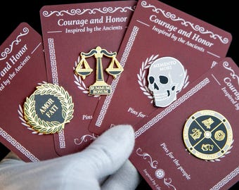 May include: A collection of pins on burgundy cards. The pins feature designs such as a scale, a skull, and a circular emblem with symbols. The cards have the text "Courage and Honor" and "Inspired by the Ancients".