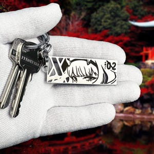May include: A silver keychain with two keys and a rectangular charm. The charm features a black and white anime-style illustration of a character's face. The keychain is held in a gloved hand.
