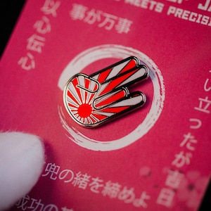 Könnte beinhalten: Ein rot-weiß-goldener Emaille-Pin in Form einer Handgeste mit einem Design der aufgehenden Sonne. Der Pin befindet sich vor einem rosa Hintergrund mit japanischem Text.