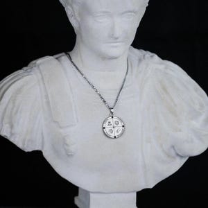 Puede incluir: Un collar con colgante plateado con un colgante circular. El colgante tiene cuatro secciones con símbolos y texto. El collar se muestra sobre un busto blanco sobre un fondo negro. La cadena es delicada.