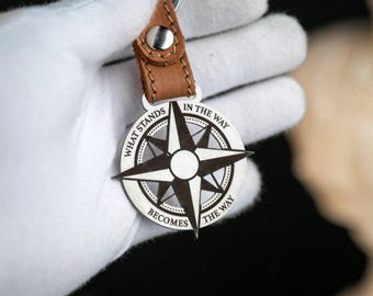 May include: A silver compass keychain with a brown leather strap. The compass design features a starburst pattern and the engraved text "WHAT STANDS IN THE WAY BECOMES THE WAY". The keychain is held by a gloved hand.