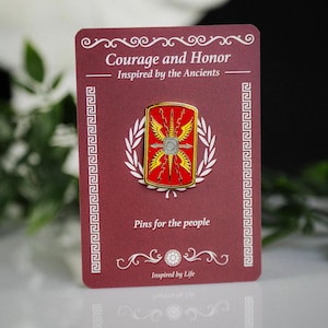May include: A burgundy card with a Roman-themed enamel pin. The pin is red and gold, with a central emblem and a white laurel wreath. Text on the card reads "Courage and Honor" and "Pins for the people".