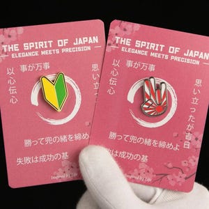 Puede incluir: Dos tarjetas rosas con texto japonés y alfileres. Un alfiler es un símbolo de conductor principiante verde y amarillo. El otro alfiler es un gesto de la mano rojo y blanco con un diseño de sol naciente. Las tarjetas tienen el texto "THE SPIRIT OF JAPAN" y "ELEGANCE MEETS PRECISION".