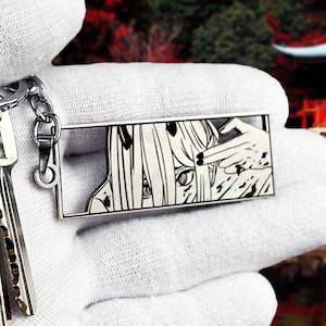 May include: A silver-colored keychain featuring a rectangular pendant with a black and white anime-style illustration. The illustration depicts a character with long hair and a hand pointing towards their eye. The keychain includes a key ring and a chain.