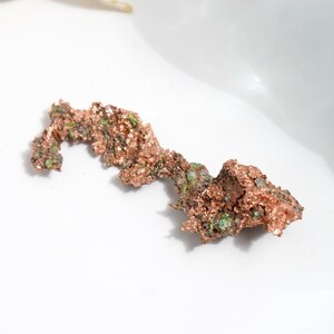 May include: A raw copper specimen with a textured, irregular shape. The copper has a reddish-brown color with patches of green and is displayed against a white background. This natural mineral sample is ideal for collectors and educational purposes.