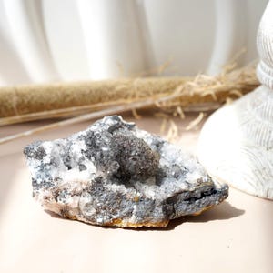 May include: A raw, natural crystal cluster with a mix of gray, white, and clear quartz crystals. The crystal formation has a rough, textured surface and sits on a light pink surface. The crystal is a natural mineral specimen.