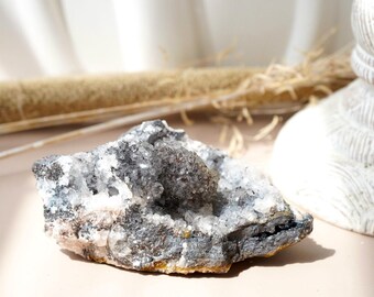May include: A raw, natural crystal cluster with a mix of gray, white, and clear quartz crystals. The crystal formation has a rough, textured surface and sits on a light pink surface. The crystal is a natural mineral specimen.