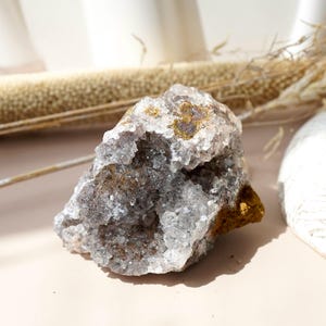 May include: A raw, uncut crystal cluster with a mix of clear, gray, and golden-brown hues. The natural mineral formation has a rough, textured surface and is displayed on a light pink surface. The crystal is a unique decorative item.