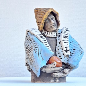 May include: A ceramic bust sculpture of a figure wearing a hooded cloak and holding a reddish-orange orb. The cloak is light blue with white accents and the hood is tan. The figure's face is serene.