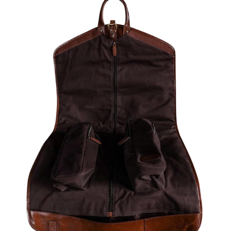 Op de afbeelding: Een bruine kledingtas met een leren handvat en afwerking. De tas is open en toont een donkerbruin interieur met twee kleine zakken. Een rits loopt in het midden en de tas is ontworpen om kleding te vervoeren.