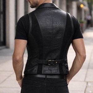 May include: A black vest featuring a textured crocodile-skin pattern on the back panels, contrasted with smooth black side panels. The garment includes a horizontal adjustable strap across the lower back with a metallic buckle and a collar.