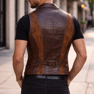May include: A brown vest featuring a combination of textured crocodile-embossed leather panels and smooth suede side sections. The garment includes a notched collar and an adjustable back strap with a metallic buckle, worn over a black t-shirt.
