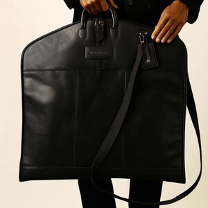 May include: A black leather garment bag with a top handle, buckle closure, and a detachable shoulder strap. The bag has a rectangular shape with a small tag attached to the strap. The brand name is embossed on the front.
