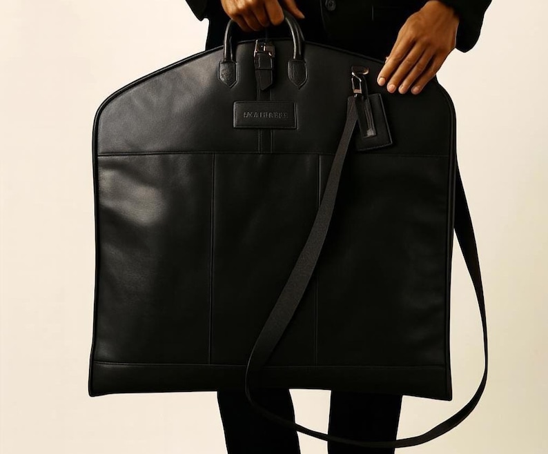 May include: A black leather garment bag with a top handle, buckle closure, and a detachable shoulder strap. The bag has a rectangular shape with a small tag attached to the strap. The brand name is embossed on the front.
