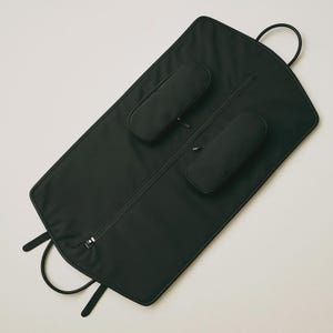 May include: Black garment bag with a full-length zipper, two padded sections, and a top handle. The bag has two small handles at the bottom and is designed to protect clothing during travel.