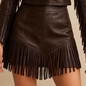 May include: A dark brown leather skirt with a fringed hemline. The skirt has a high waist and a tailored fit. The fringe is long and hangs down from the hemline. The skirt is part of a matching leather outfit.