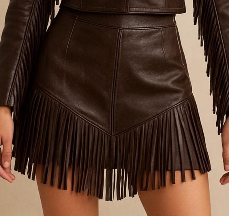 May include: A dark brown leather skirt with a fringed hemline. The skirt has a high waist and a tailored fit. The fringe is long and hangs down from the hemline. The skirt is part of a matching leather outfit.