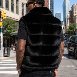 May include: A black fur vest with a horizontal quilted design. The vest has a high collar and is worn over a black t-shirt. The person is wearing black jeans and sunglasses. The background shows a city street with buildings and cars.