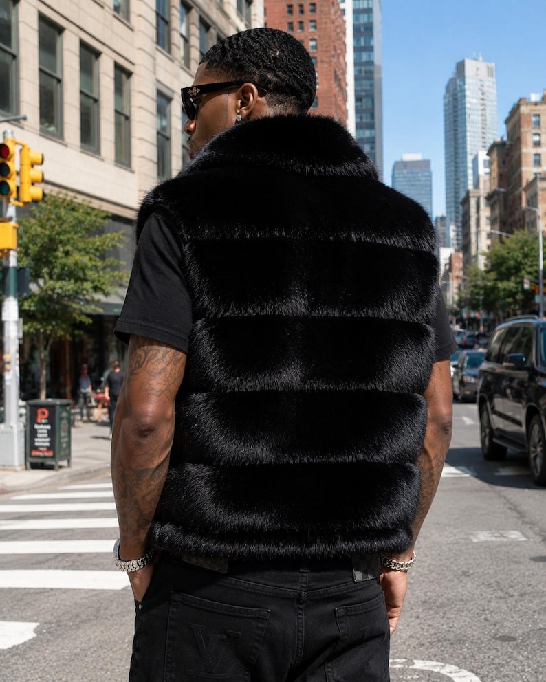 May include: A black fur vest with a horizontal quilted design. The vest has a high collar and is worn over a black t-shirt. The person is wearing black jeans and sunglasses. The background shows a city street with buildings and cars.
