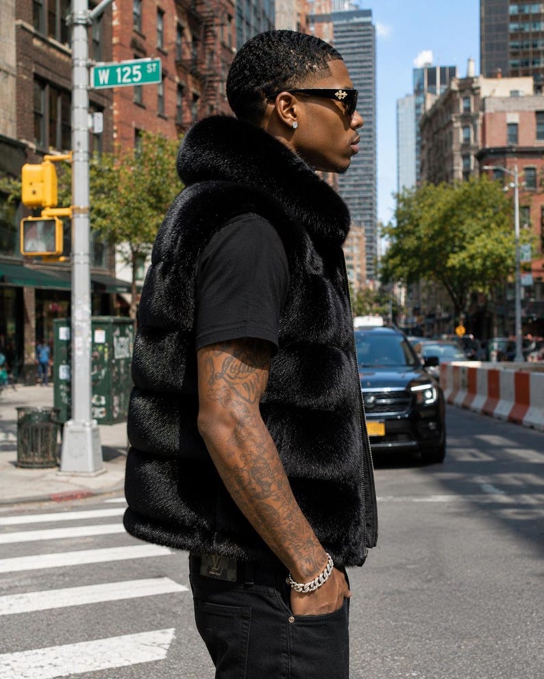 May include: A black fur vest with a high collar. The vest is worn over a black t-shirt and paired with black jeans. The person is wearing sunglasses and a silver bracelet. The background shows a city street with a street sign that reads W 125 ST.