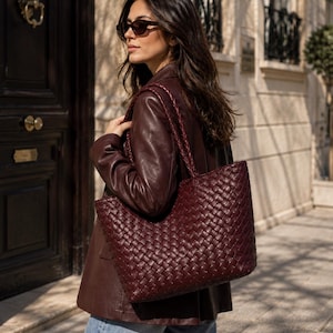 Puede incluir: Un bolso tote de color burdeos intenso con un intrincado patrón de cuero trenzado y dos correas de hombro trenzadas. El bolso tiene una forma rectangular con una abertura ancha y un acabado rojo oscuro uniforme en toda la superficie.