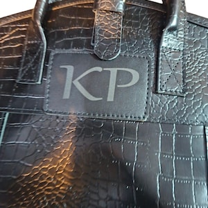 May include: A black handbag with a textured crocodile-embossed pattern featuring a rectangular patch on the front center with the letters KP embossed in a serif font. The bag includes two top handles and a central strap with a gold-tone buckle.
