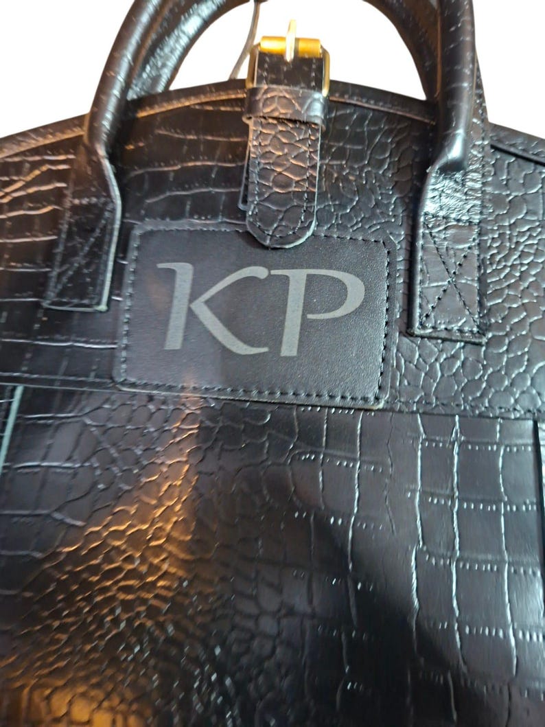 May include: A black handbag with a textured crocodile-embossed pattern featuring a rectangular patch on the front center with the letters KP embossed in a serif font. The bag includes two top handles and a central strap with a gold-tone buckle.