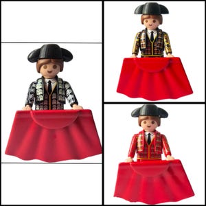 May include: Three plastic toy figures dressed in traditional matador attire, each wearing a black montera hat, a white shirt with a black tie, and a detailed jacket. Each figure holds a large, solid red cape in front of their body.