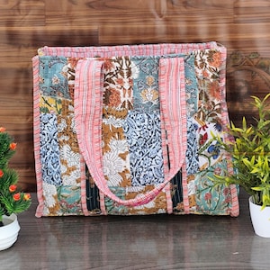 以下が含まれることがあります： 青、茶色、白の色合いの花柄模様のパッチワークトートバッグ。バッグにはピンクのストライプのハンドルとトリムが付いています。バッグは長方形で、キルティング生地で作られているようです。