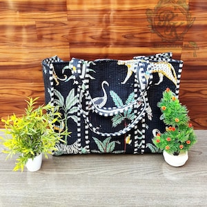 May include: Black tote bag with a jungle print featuring zebras, leopards, and tropical plants. The bag has white and black striped accents and sturdy handles. The bag is sitting on a wooden surface.
