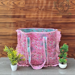 Könnte beinhalten: Eine pinkfarbene Tragetasche mit floralem Muster und Rüschenkanten. Die Tasche hat türkis- und pinkfarbene gestreifte Griffe und Verzierungen. Die Tasche steht auf einer grauen Oberfläche mit kleinen Topfpflanzen auf beiden Seiten.