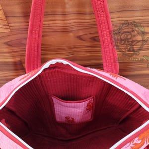 May include: Pink tote bag with red accents and a white zipper. The bag features a red interior and a small pocket. The exterior has an orange lobster design. The handles are red and quilted.