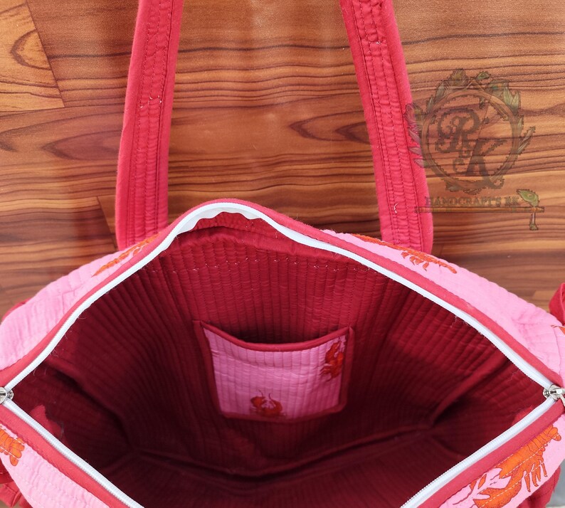 May include: Pink tote bag with red accents and a white zipper. The bag features a red interior and a small pocket. The exterior has an orange lobster design. The handles are red and quilted.