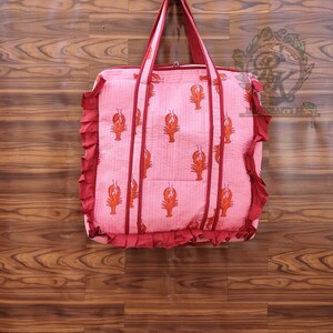 May include: A pink tote bag with a repeating pattern of orange lobsters. The bag has red trim, handles, and a ruffled edge. The bag is made of a quilted fabric and is designed for carrying items.