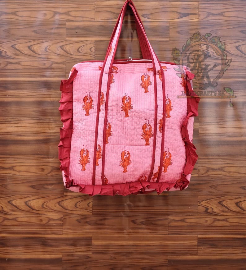 May include: A pink tote bag with a repeating pattern of orange lobsters. The bag has red trim, handles, and a ruffled edge. The bag is made of a quilted fabric and is designed for carrying items.