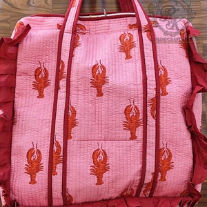 May include: Pink quilted tote bag with a repeating pattern of orange lobsters. The bag has dark red trim, handles, and ruffled edges. The bag is designed for carrying items.