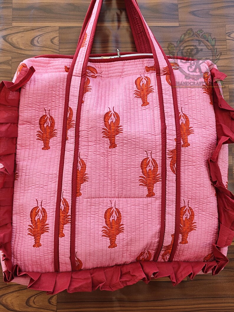 May include: Pink quilted tote bag with a repeating pattern of orange lobsters. The bag has dark red trim, handles, and ruffled edges. The bag is designed for carrying items.