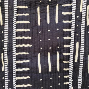 May include: Close-up of a black and white patterned textile. The design features vertical stripes with alternating patterns of white dashes, lines, and dots. The fabric has a quilted texture, with visible stitching throughout.