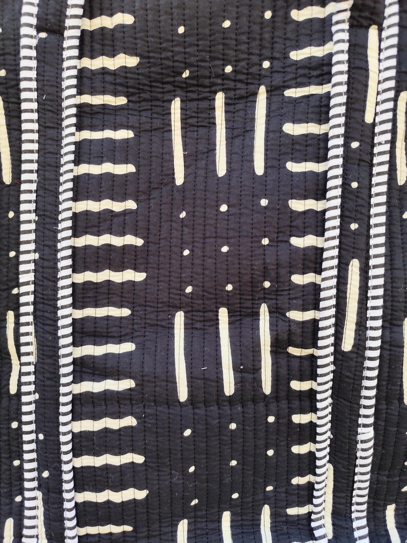 May include: Close-up of a black and white patterned textile. The design features vertical stripes with alternating patterns of white dashes, lines, and dots. The fabric has a quilted texture, with visible stitching throughout.