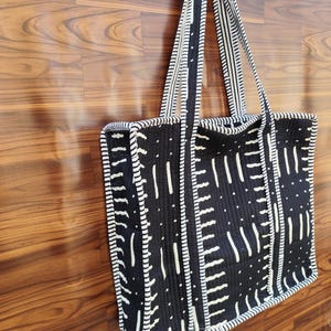 May include: A black and white tote bag with a geometric pattern. The bag features a repeating design of vertical and horizontal white lines on a black background. The handles and edges are also white and black striped.