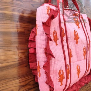 May include: Pink quilted tote bag with a repeating orange lobster print. The bag has red trim, ruffled edges, and matching red handles. The bag is made of a soft, quilted material.