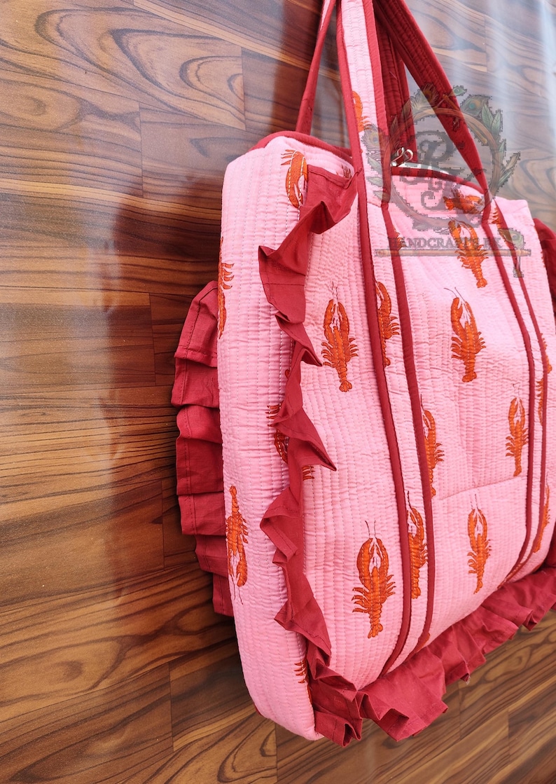 May include: Pink quilted tote bag with a repeating orange lobster print. The bag has red trim, ruffled edges, and matching red handles. The bag is made of a soft, quilted material.